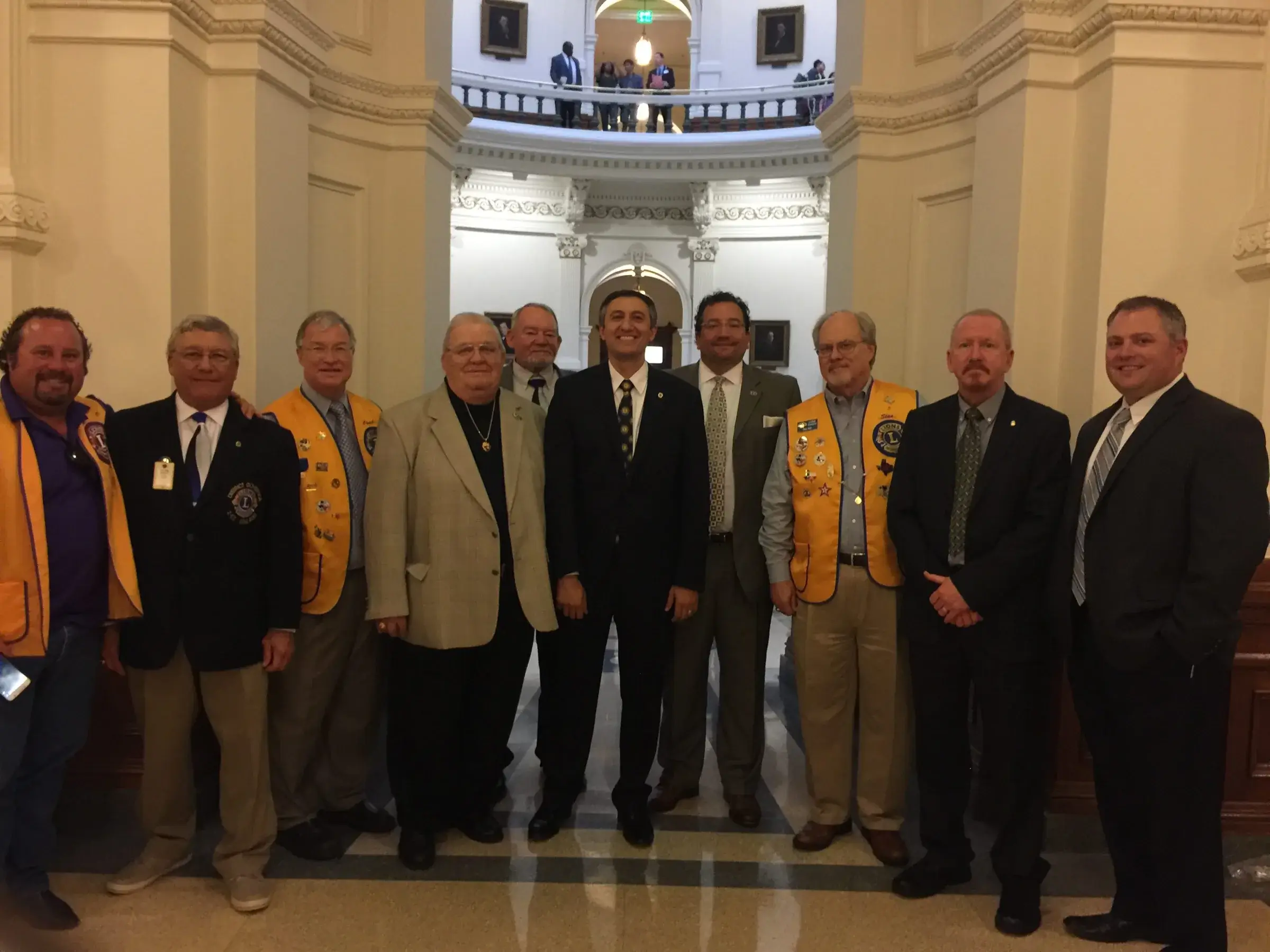 Eleven men stand together in the marble rotunda of the Texas State Capitol, several wearing gold Lions vests heavy with pins and one in a purple and gold vest, alongside others in business suits. Framed portraits and a balustraded gallery are visible above them.